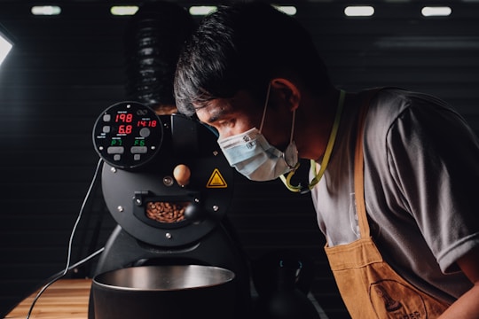 A person wearing a mask and an apron is closely observing a coffee roasting machine. The machine's display shows various numbers related to temperature and time, and coffee beans are visible inside. The environment suggests an industrial or professional setting with dim lighting.