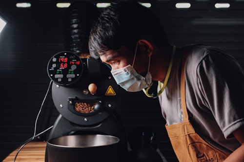 A person wearing a mask and an apron is closely observing a coffee roasting machine. The machine's display shows various numbers related to temperature and time, and coffee beans are visible inside. The environment suggests an industrial or professional setting with dim lighting.