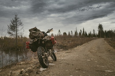 A rugged motorcycle loaded with camping gear is parked on a dirt road surrounded by a sparse, forested landscape. The sky is overcast with dark, brooding clouds, adding a sense of adventure and isolation. The path stretches into the distance beside a narrow body of water, with leafless trees scattered around.