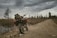 A rugged motorcycle loaded with camping gear is parked on a dirt road surrounded by a sparse, forested landscape. The sky is overcast with dark, brooding clouds, adding a sense of adventure and isolation. The path stretches into the distance beside a narrow body of water, with leafless trees scattered around.