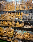 A display of intricately carved wooden statues and sculptures, featuring various deities, animals, and decorative objects. Some figurines are polished wood, while others are painted or have metallic finishes. The collection includes multiple rows of figurines of different sizes, some with small Indian flags beside them. Behind the figures are circular decorative plates depicting famous Indian landmarks.