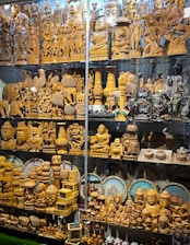 A display of intricately carved wooden statues and sculptures, featuring various deities, animals, and decorative objects. Some figurines are polished wood, while others are painted or have metallic finishes. The collection includes multiple rows of figurines of different sizes, some with small Indian flags beside them. Behind the figures are circular decorative plates depicting famous Indian landmarks.