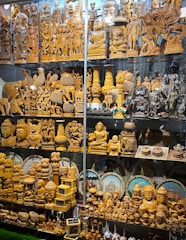 A display of intricately carved wooden statues and sculptures, featuring various deities, animals, and decorative objects. Some figurines are polished wood, while others are painted or have metallic finishes. The collection includes multiple rows of figurines of different sizes, some with small Indian flags beside them. Behind the figures are circular decorative plates depicting famous Indian landmarks.
