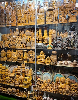 A display of intricately carved wooden statues and sculptures, featuring various deities, animals, and decorative objects. Some figurines are polished wood, while others are painted or have metallic finishes. The collection includes multiple rows of figurines of different sizes, some with small Indian flags beside them. Behind the figures are circular decorative plates depicting famous Indian landmarks.