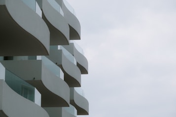 The image features a modern building with a series of uniquely shaped balconies. Each balcony has a curved design, creating an artistic and wave-like appearance. The exterior of the building is made of light-colored materials, and the sky appears overcast, enhancing the soft and minimalistic look.
