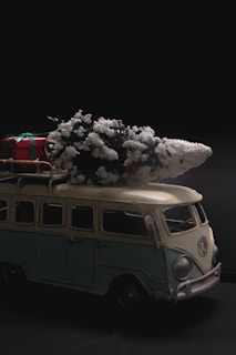 A miniature vintage van model, colored in pale green and cream, with a snow-frosted Christmas tree and a wrapped gift on its roof. The scene conveys a festive and nostalgic atmosphere against a dark background.