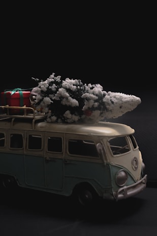 A miniature vintage van model, colored in pale green and cream, with a snow-frosted Christmas tree and a wrapped gift on its roof. The scene conveys a festive and nostalgic atmosphere against a dark background.