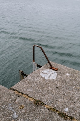 A concrete surface with rusted metal railing overlooking a body of water. The surface has visible patches of wear, and some greenery grows in the cracks.