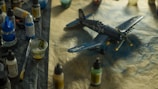 A detailed model airplane is surrounded by various painting supplies, including paints, brushes, and containers. The plane is a dark blue color with military-style markings and is resting on a textured surface.