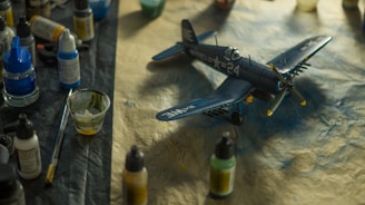 A detailed model airplane is surrounded by various painting supplies, including paints, brushes, and containers. The plane is a dark blue color with military-style markings and is resting on a textured surface.