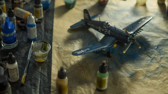 A detailed model airplane is surrounded by various painting supplies, including paints, brushes, and containers. The plane is a dark blue color with military-style markings and is resting on a textured surface.