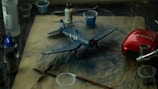 A detailed scene of a model airplane surrounded by various painting supplies and tools on a paper-covered surface. The model is a navy blue color with white markings and numbers. There are brushes, cups, and paint bottles scattered around, creating a workspace atmosphere. A noticeable red object, possibly an airbrush or paint sprayer, is also present.