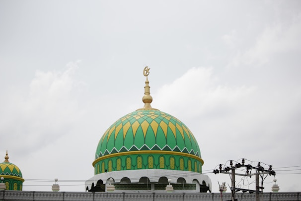 A large, ornate green and yellow dome with intricate geometric patterns and an Islamic crescent symbol on top. The dome is part of a mosque structure, featuring smaller domes and decorative arches. The sky is overcast, and there are power lines in the foreground.