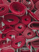 Several rolled-up red carpets are stacked closely together. The edges of the carpets fray slightly, and the rolls are tightly wound, showcasing different patterns and layers within the rolls.