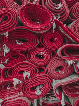 Several rolled-up red carpets are stacked closely together. The edges of the carpets fray slightly, and the rolls are tightly wound, showcasing different patterns and layers within the rolls.