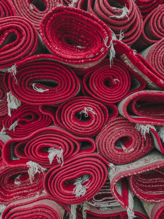 Several rolled-up red carpets are stacked closely together. The edges of the carpets fray slightly, and the rolls are tightly wound, showcasing different patterns and layers within the rolls.