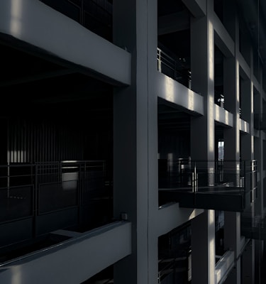 The image features a dark interior space with structural beams and shadows creating a geometric pattern. The lighting highlights the grid-like architecture, giving depth to the expansive framework. There are multiple railing sections along the platforms and walkways.