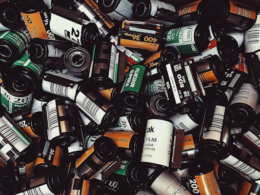 A pile of various film rolls, including brands like Kodak and Fujicolor, with visible labels indicating different speeds such as 200 and 400 ISO. The film cases are predominantly cylindrical with a mix of colors including black, white, orange, and green.