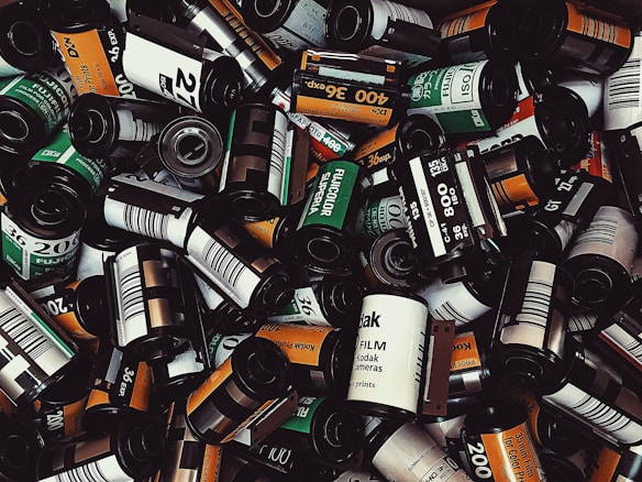 A pile of various film rolls, including brands like Kodak and Fujicolor, with visible labels indicating different speeds such as 200 and 400 ISO. The film cases are predominantly cylindrical with a mix of colors including black, white, orange, and green.