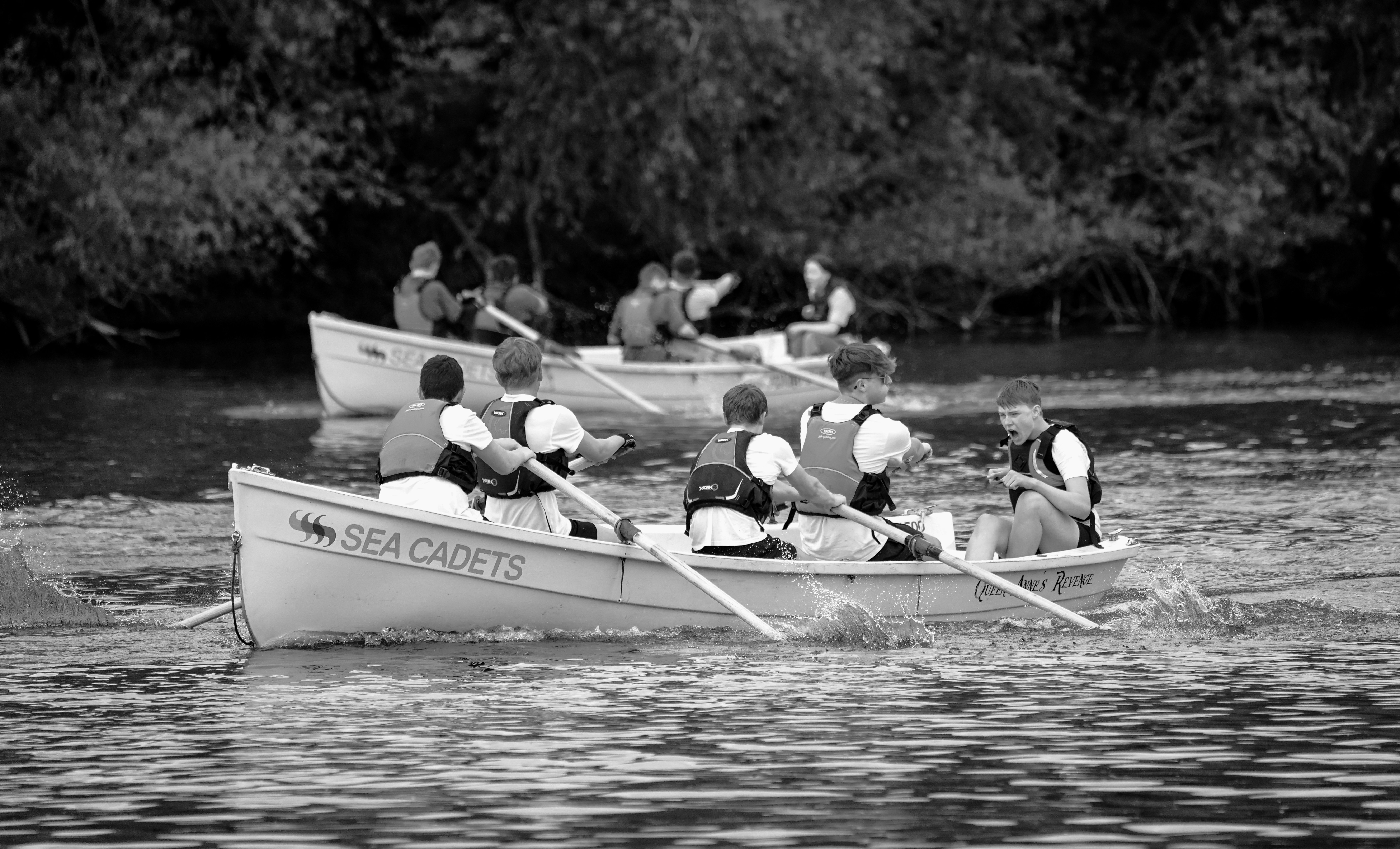 A black and white photograph of Sea Cadets rowing boats on water. The image shows two white rowing boats with crews of young people wearing life jackets, actively rowing with oars.