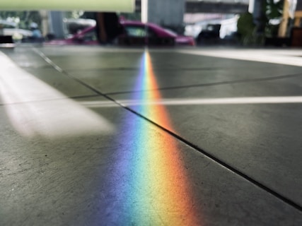 A vibrant spectrum of rainbow colors is distinctly visible on a tiled floor surface, with hues ranging from red to violet, creating a striking visual effect. The surrounding area appears to be indoors or semi-outdoor, with blurred elements of parked vehicles and greenery in the background.