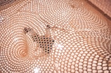A detailed pattern of woven mesh creates a captivating visual, highlighting an intricate geometric design. A person is partially visible beneath the mesh, adding depth and interest to the scene, with light reflecting off the surface.
