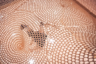 A detailed pattern of woven mesh creates a captivating visual, highlighting an intricate geometric design. A person is partially visible beneath the mesh, adding depth and interest to the scene, with light reflecting off the surface.