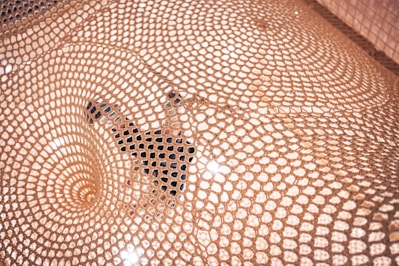 A detailed pattern of woven mesh creates a captivating visual, highlighting an intricate geometric design. A person is partially visible beneath the mesh, adding depth and interest to the scene, with light reflecting off the surface.