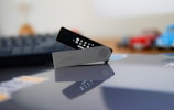 A Ledger hardware wallet is positioned on a reflective surface, with its screen displaying small icons and text. The device is angled open, showing its brand name on the front panel. In the blurred background, a computer keyboard and some indistinct objects in various colors are visible.