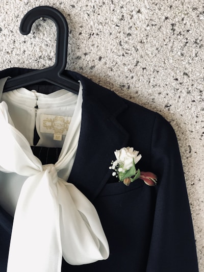 A dark suit jacket hangs on a black hanger against a textured speckled wall. The jacket features a prominent white boutonnière with a white flower and green leaves in the pocket. Draped beneath the jacket lapel, a white blouse with a large bow adds a striking contrast.