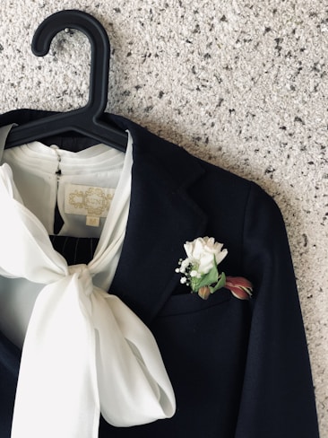 A dark suit jacket hangs on a black hanger against a textured speckled wall. The jacket features a prominent white boutonni&egrave;re with a white flower and green leaves in the pocket. Draped beneath the jacket lapel, a white blouse with a large bow adds a striking contrast.