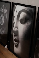 A detailed close-up photograph of a serene, large-scale Buddha face sculpture. The artwork is positioned in a tilted frame against a muted wall, capturing the serene expression. The smooth texture and subtle grain of the sculpture are highlighted by soft lighting.