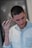 A person with short hair is using an electric clipper to trim their hair. They have a small tattoo near the hairline and a tattoo visible on the neck. The person is wearing a light denim shirt and has a ring on one finger. The background is blurred, emphasizing the focus on the person.