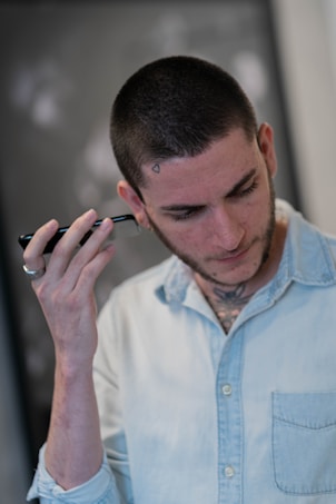 A person with short hair is using an electric clipper to trim their hair. They have a small tattoo near the hairline and a tattoo visible on the neck. The person is wearing a light denim shirt and has a ring on one finger. The background is blurred, emphasizing the focus on the person.