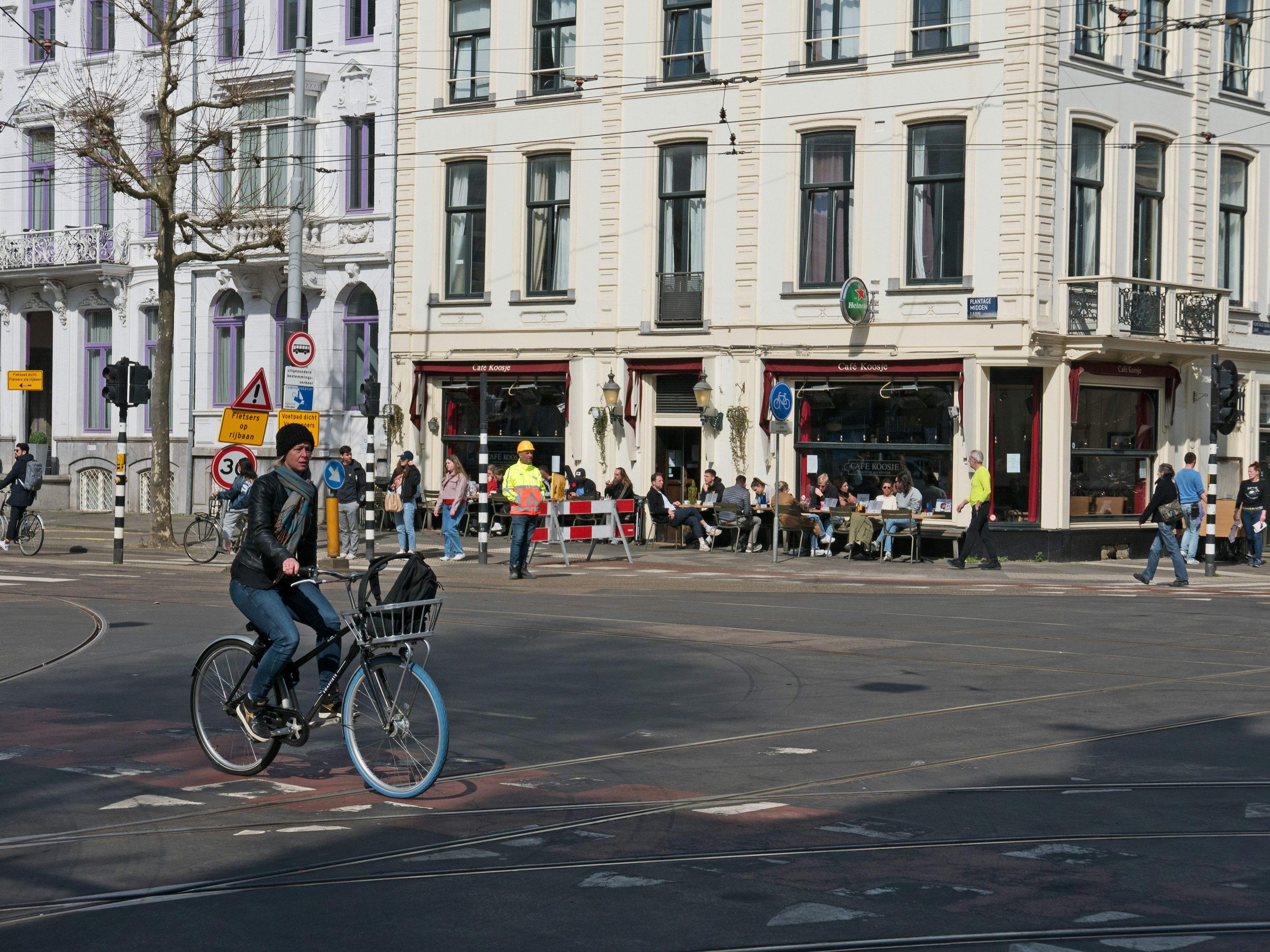 Woman is biking over the crossing of the streets Plantage Middenlaan / Plantage Kerklaan - in Amsterdam city on a sunny day in Spring. A pub with terrace is located here at the streetcorner with a nice view over the street and people in sunlight. Free street photo Amsterdam people by Fons Heijnsbroek, Netherlands 28 April 2022 // Rechtenvrije, gratis download foto in hoge resolutie van straten in Amsterdam; een fietser rijdt op het kruispunt van de Plantage Kerklaan en de Plantage Middenlaan met mensen die in de zon op het terras zitten. Foto, Fons Heijnsbroek - straatfotografie, Nederland.