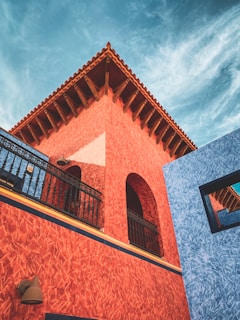 A vibrant architectural scene featuring a building with textured, red-orange walls and a wooden roof. The structure showcases detailed elements such as a black wrought iron balcony and a vivid blue adjacent wall. The sky above is a striking blue with wispy clouds, creating a dynamic backdrop.