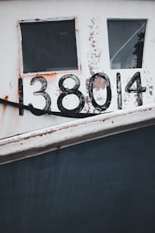 A close-up view of a weathered boat side with visible rust and paint peeling. Two rectangular windows are positioned above the number '138014,' which is painted in large, bold black numbers. The surface shows signs of age and exposure to elements.
