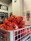A collection of bright orange soccer balls with black patterns are piled inside a metal basket. The background is slightly blurred, showing part of a store interior with shelves.