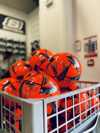 A collection of bright orange soccer balls with black patterns are piled inside a metal basket. The background is slightly blurred, showing part of a store interior with shelves.