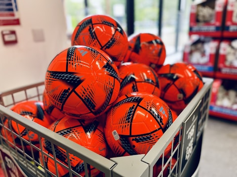 A metal bin is filled with several bright orange soccer balls featuring black graphic patterns. The balls are neatly arranged in a store setting, with shelves visible in the background.