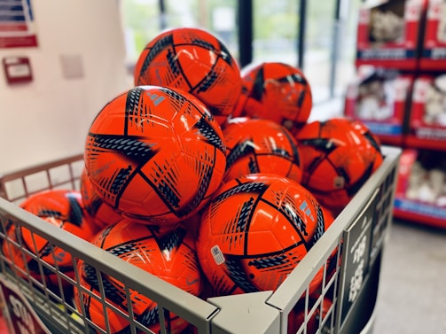 A metal bin is filled with several bright orange soccer balls featuring black graphic patterns. The balls are neatly arranged in a store setting, with shelves visible in the background.