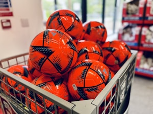 A metal bin is filled with several bright orange soccer balls featuring black graphic patterns. The balls are neatly arranged in a store setting, with shelves visible in the background.
