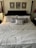 A neatly made bed with a high, dark wooden headboard features multiple pillows including a central striped decorative pillow. Above the headboard, three framed artworks are symmetrically arranged on a light-colored wall. The bedspread is light in color, and there is a small nightstand to the side with a lamp and a device, possibly an alarm clock.
