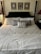 A neatly made bed with a high, dark wooden headboard features multiple pillows including a central striped decorative pillow. Above the headboard, three framed artworks are symmetrically arranged on a light-colored wall. The bedspread is light in color, and there is a small nightstand to the side with a lamp and a device, possibly an alarm clock.