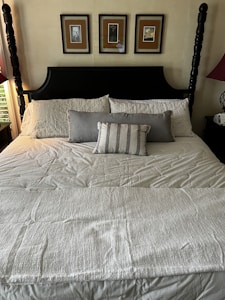 A neatly made bed with a high, dark wooden headboard features multiple pillows including a central striped decorative pillow. Above the headboard, three framed artworks are symmetrically arranged on a light-colored wall. The bedspread is light in color, and there is a small nightstand to the side with a lamp and a device, possibly an alarm clock.