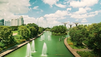 A lush green park with various types of trees and plants surrounding a serene body of water. In the background, a large Ferris wheel and several futuristic tree-like structures rise against a backdrop of modern skyscrapers. The sky is bright with fluffy clouds, and there are fountains creating splashes in the water.