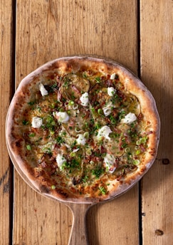 A freshly baked pizza with a crispy, golden-brown crust topped with sliced zucchini, dollops of creamy cheese, and a sprinkling of green herbs over a wooden paddle on a rustic wooden table.