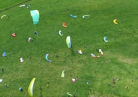 Aerial view of a grassy field dotted with colorful paragliders and people preparing for paragliding. The scene is lively with various paragliders in shades of blue, green, red, yellow, and orange scattered across the lush green grass. Several individuals are seen scattered around, likely adjusting equipment or preparing for takeoff.