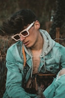 A person wearing white-rimmed sunglasses and a denim jacket layered over a gray hoodie is seated in an outdoor setting. They are carrying a designer bag with a distinct monogram pattern. Their hair is styled voluminously, and there is a relaxed yet introspective demeanor.