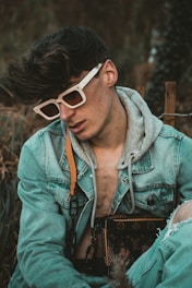 A person wearing white-rimmed sunglasses and a denim jacket layered over a gray hoodie is seated in an outdoor setting. They are carrying a designer bag with a distinct monogram pattern. Their hair is styled voluminously, and there is a relaxed yet introspective demeanor.